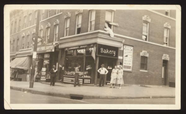 Front shows an American street scene with "Langenbein's Bakery" and "Paint Supply" shop signs. Several people standing in front. Appears to be the family's American business. The annotation suggests these were sent to Germany to show relatives the family's shop.