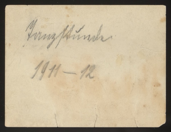 ANNOTATION ON PHOTO (back, handwritten): German: "Tanzstunde. 1911-12" English: "Dance class/lesson. 1911-12" Description: Front shows a large group of approximately 20 young people at a formal dance or social event, some holding gingerbread hearts. Taken in a room with landscape paintings on the walls. The group appears to be from the 1911-1912 winter dance season.