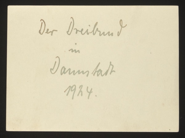 ANNOTATION ON PHOTO (back, handwritten): German: "Der Dreibund in Darmstadt 1924." English: "The Triple Alliance in Darmstadt 1924." Description: Front shows three children sitting on a garden bench — two girls and a boy (or toddler). The playful title "Der Dreibund" (The Triple Alliance) is a humorous reference to the political Triple Alliance, applied here to the three children. Photographed in Darmstadt, 1924.
