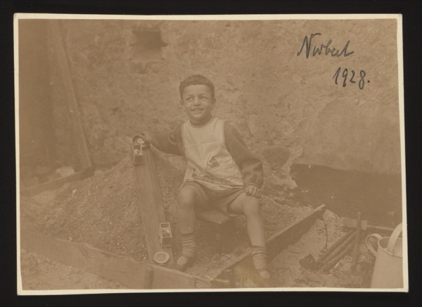 ANNOTATION ON PHOTO (front, top right, handwritten in blue ink): "Norbert 1928." Description: Boy of approximately 5-6 years old sitting in what appears to be a sandpit or construction area, playing with toys including a toy train set. Identified as Norbert, photographed in 1928.