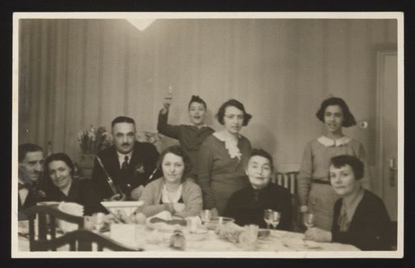 Front shows a family dinner gathering with 9 people around a table (6 adults seated, 2 young women and a boy standing behind). Glasses and dishes on the table suggest a celebration. Location: Munich, February 1932.